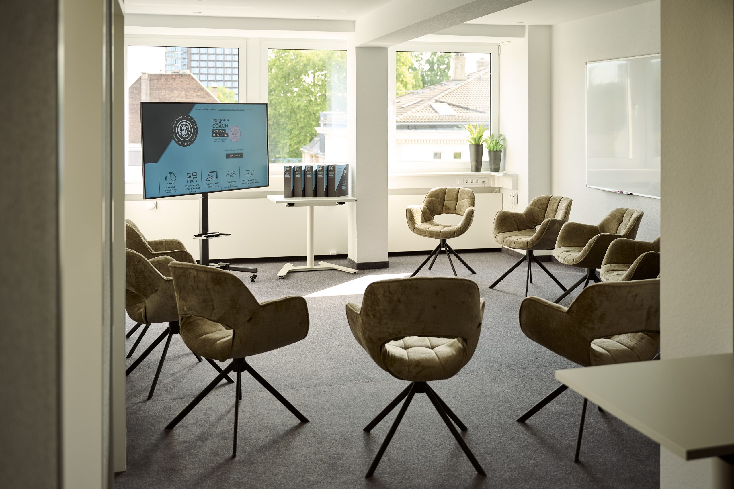 Ein heller Raum mit bequemen Stühlen, links steht ein Fernseher mit einem Präsentationschart für die Business-Life Coach Ausbildung
