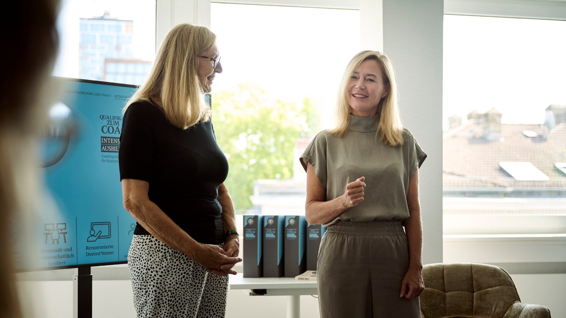 2 Frauen in legerer Businesskleidung, links Angelika Erhardt, rechts Daniela Elsner, Daniela spricht zu jemandem außerhalb des Bildes, Angelika schaut Daniela interessiert an